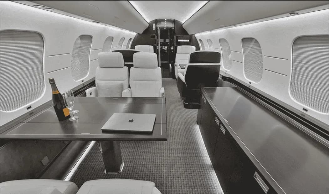 A private jet cabin interior with light-colored seats, a table with a laptop and champagne, and dark wood accents.