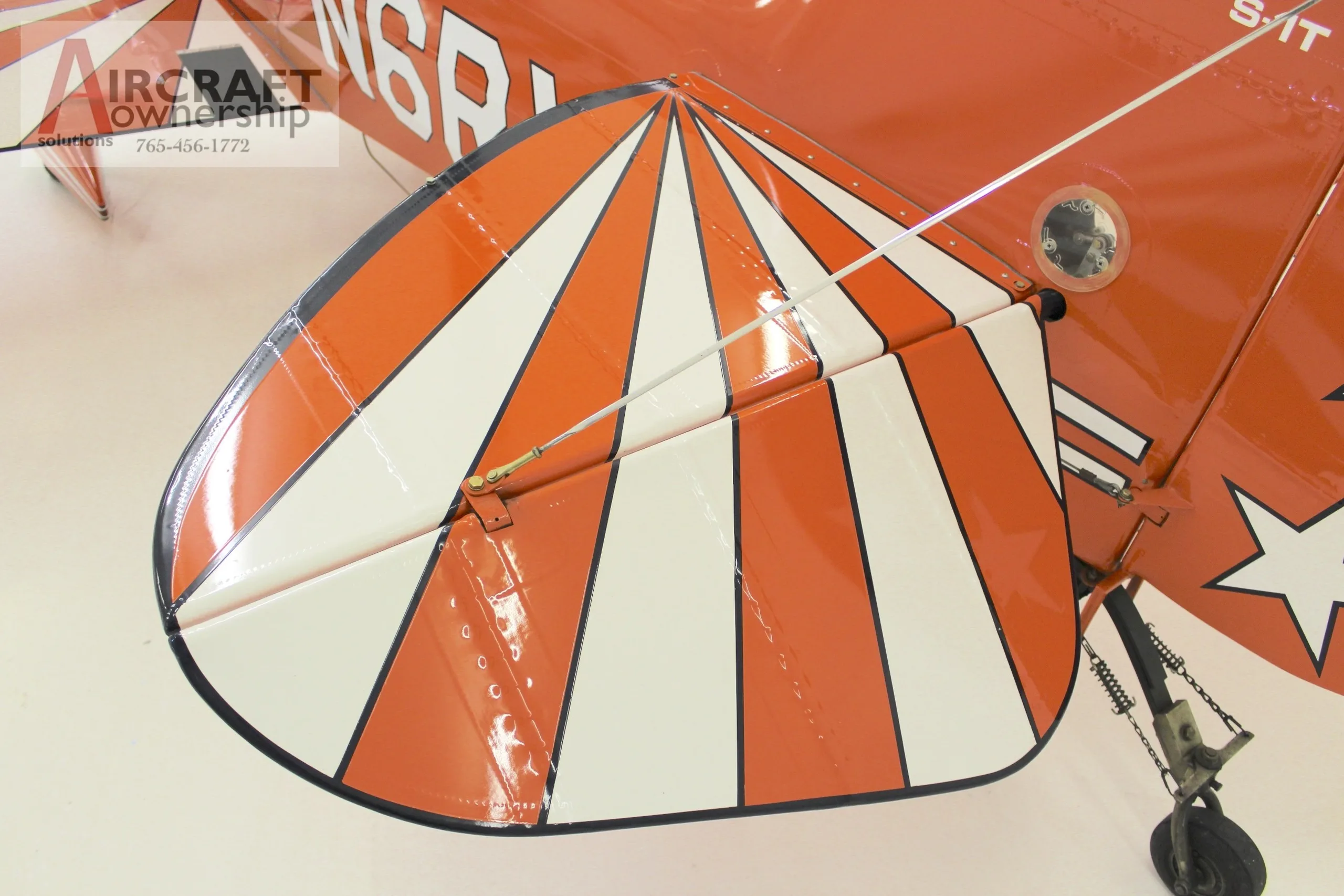 An overhead close-up view of the horizontal stabilizer of an orange Pitts S-1T biplane, featuring a white and orange sunburst pattern.