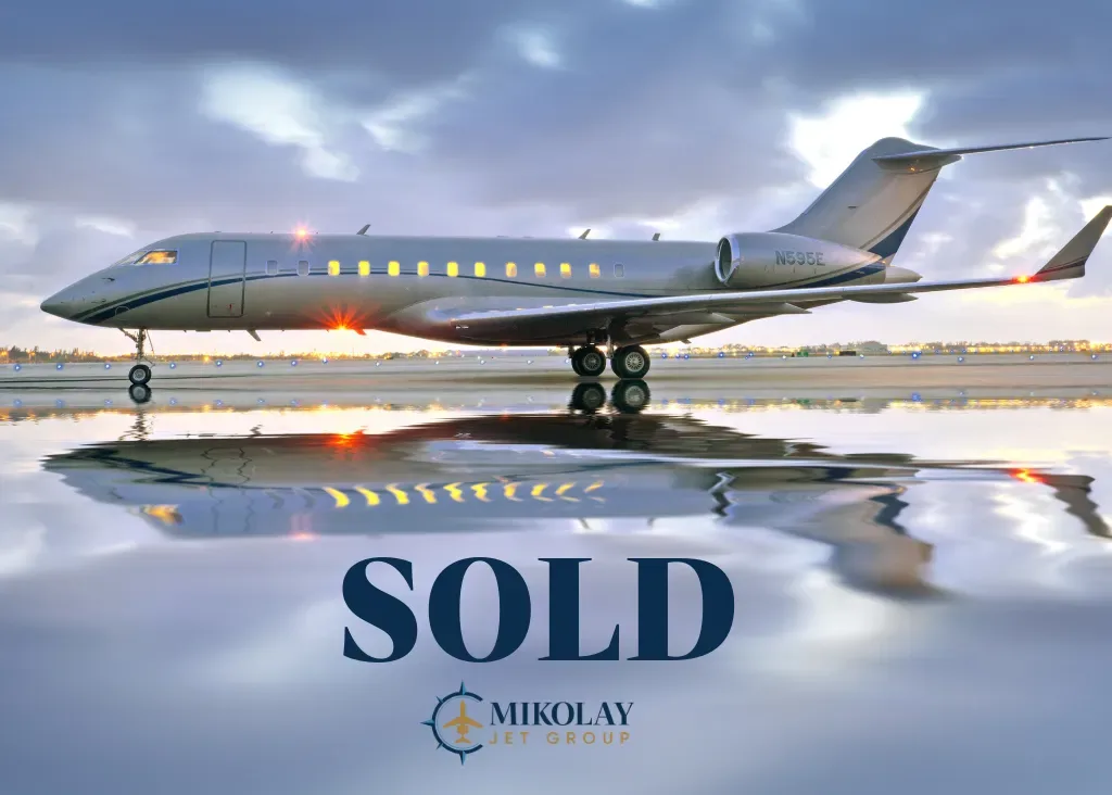 A silver Bombardier Global Express private jet (N99BE) with its lights on, reflected on a wet runway at dusk, with 'SOLD' overlaid.