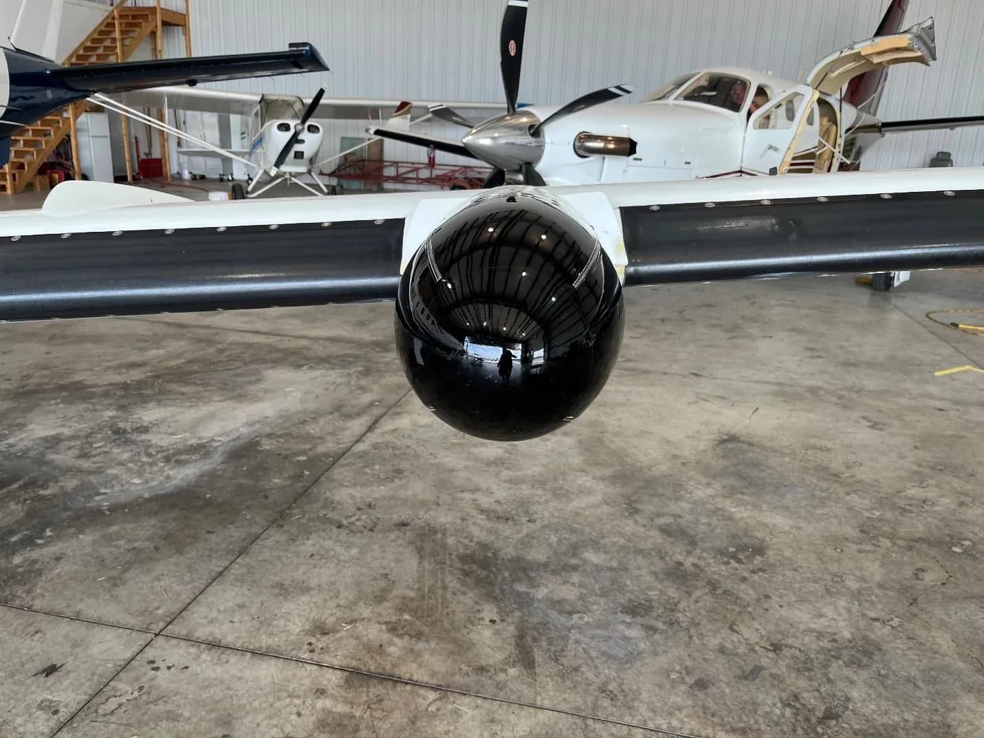 A close-up view of a black pod on the wing of a white and blue aircraft inside a hangar.