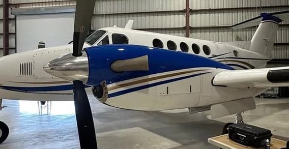 A white twin-turboprop aircraft with blue and gold stripes on the fuselage and engine nacelles, parked inside a hangar, viewed from the right side.