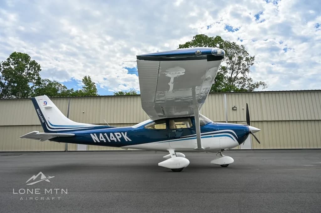 A side view of a blue and white Cessna Skylane N414PK with its wing flaps extended, parked on an asphalt ramp under a cloudy sky.