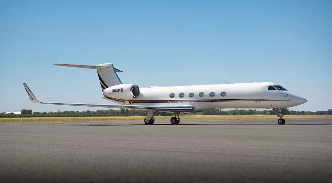 A white Gulfstream G550 business jet with red and black tail stripes, registration N631VR, parked on an airfield under a clear blue sky.