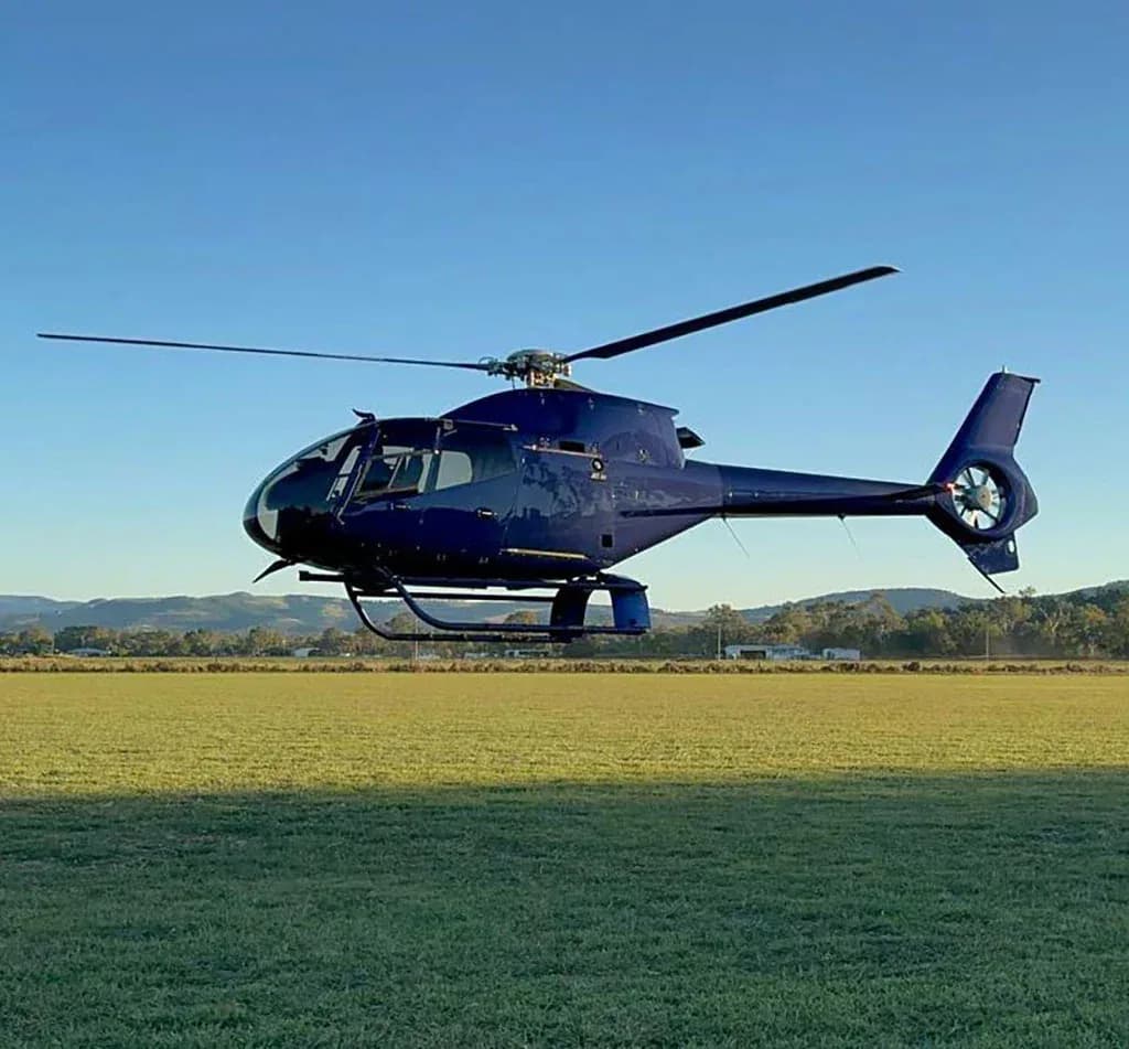 A dark blue helicopter hovers low over a green field with hills in the background under a clear blue sky.