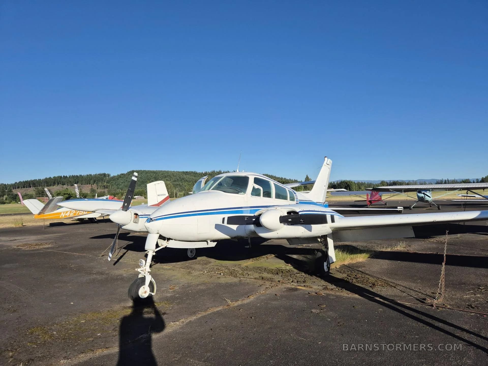 A white twin-engine aircraft with blue stripes and registration N1046Q, seen from the front-right, parked on an asphalt ramp under a clear blue sky.