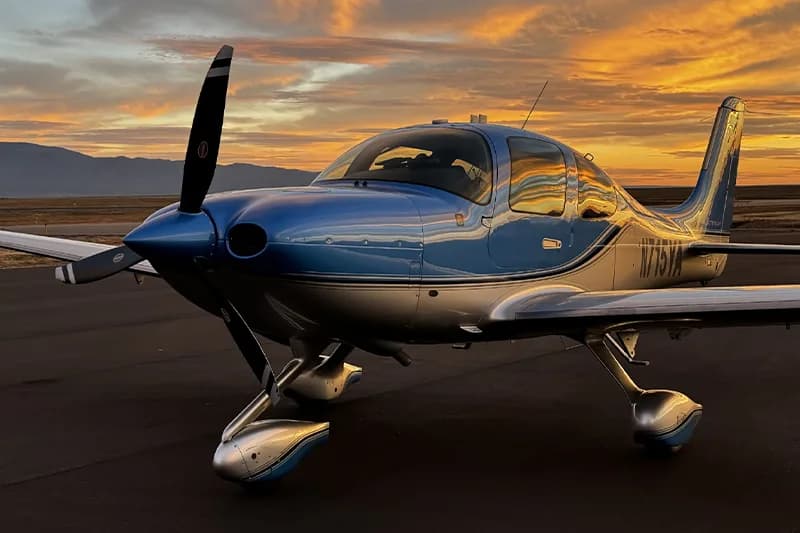 A blue and silver Cirrus aircraft parked on a runway at sunset.