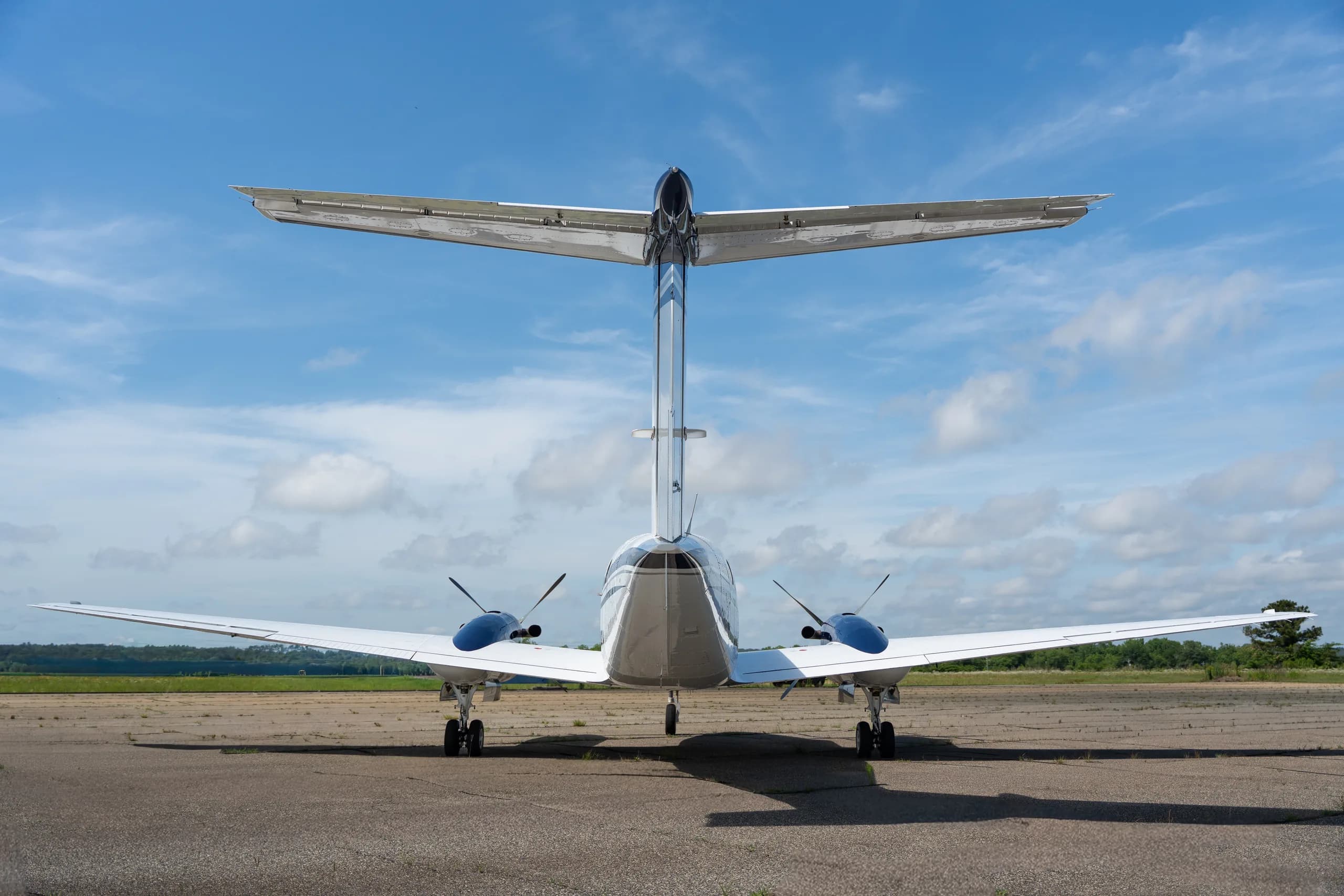 A rear view of a white and blue Beechcraft King Air twin-turboprop aircraft with a T-tail on a tarmac.