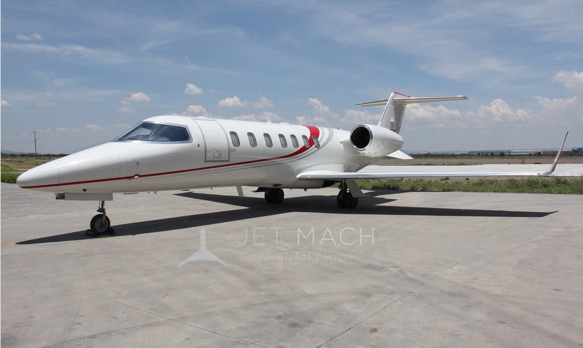 A white Learjet 75 with red and grey stripes parked on a concrete tarmac under a partly cloudy sky.