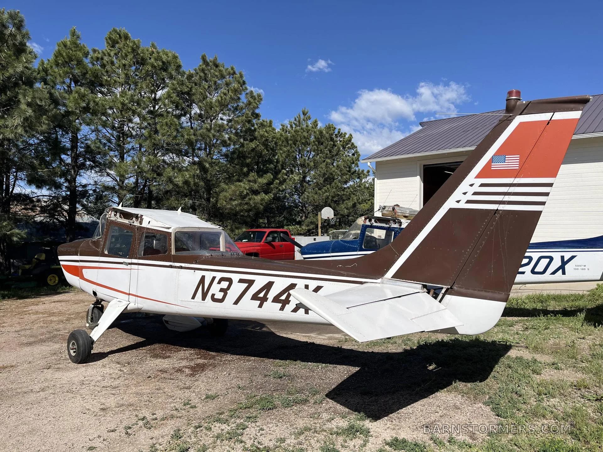 The rear-side view of a brown and white single-engine propeller aircraft (N3744X) parked on dirt next to a hangar.