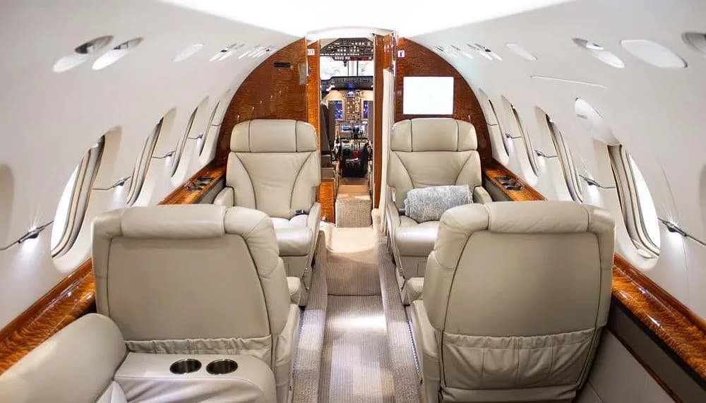 A view down the aisle of a private jet cabin, showing multiple beige leather seats and wood paneling, looking towards the cockpit.