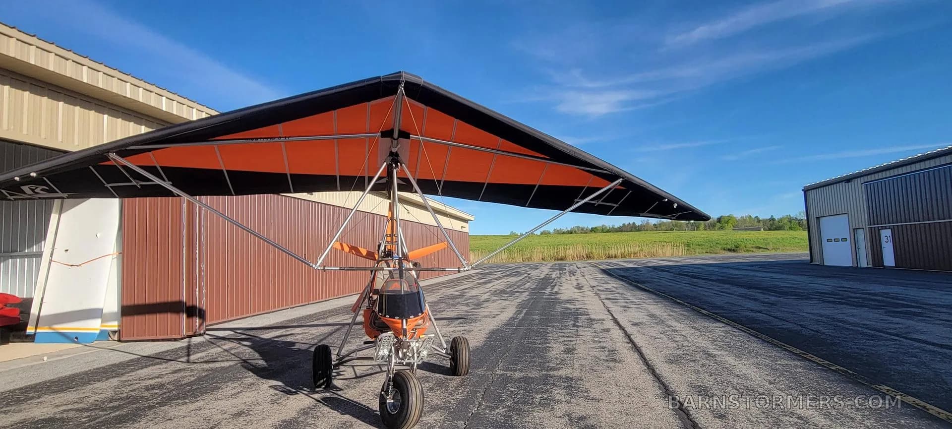 A front view of an orange and black weight-shift control ultralight aircraft on an airfield with hangars in the background.