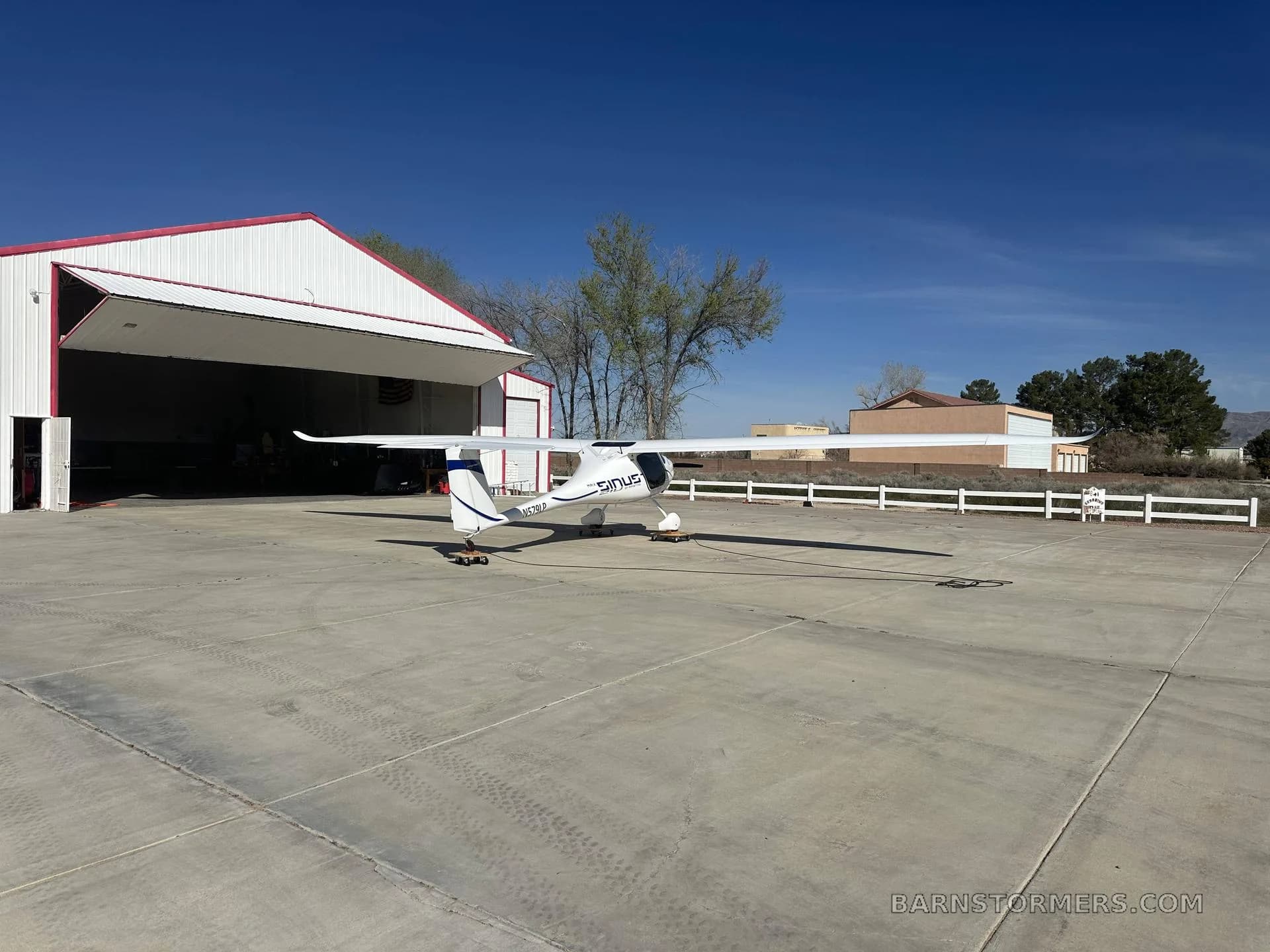 A white Pipistrel Sinus aircraft with blue accents is viewed from the rear-left, parked on an apron in front of a hangar.