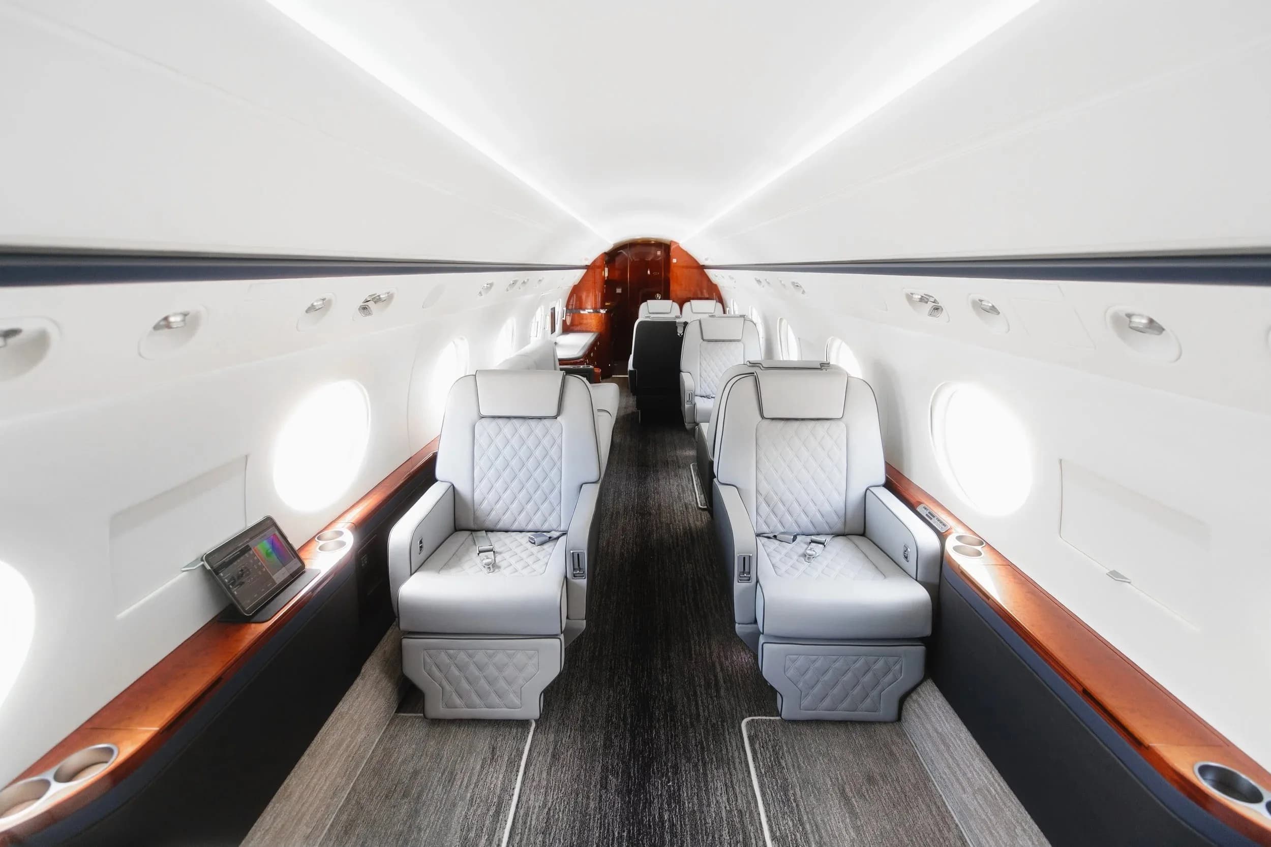 The luxurious cabin interior of a business jet, featuring light grey quilted leather seats and dark wood accents.