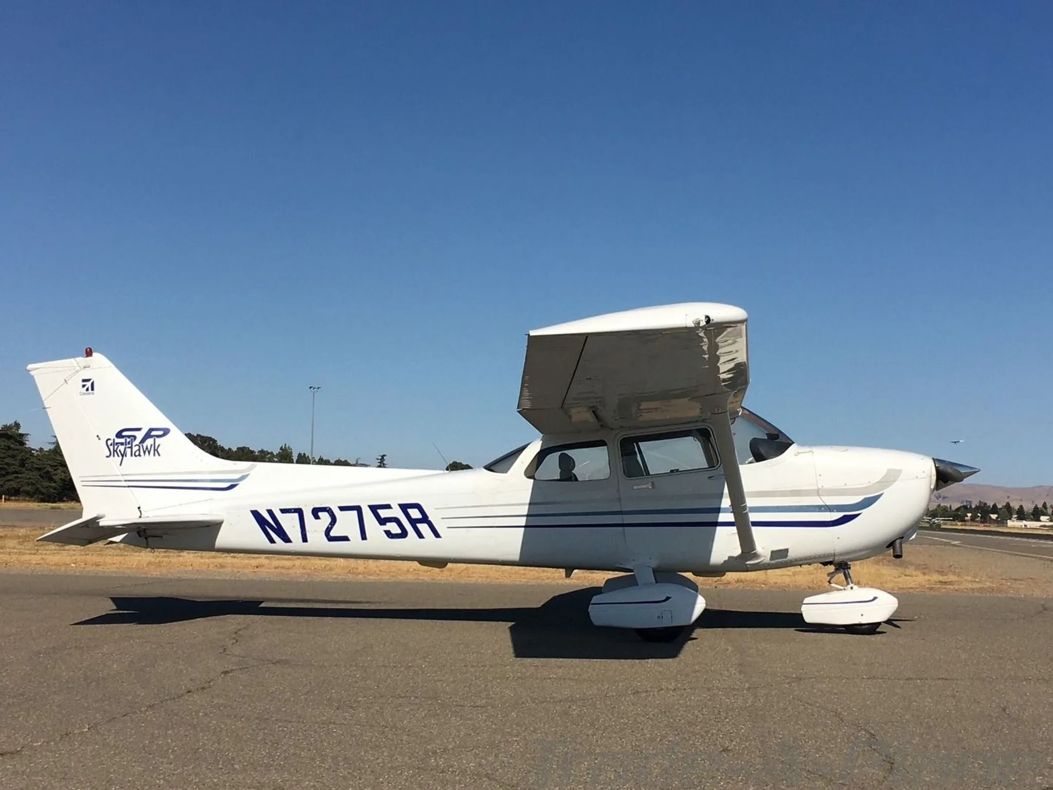 A white Cessna 172SP Skyhawk with blue stripes, registration N7275R, parked on an asphalt ramp under a clear blue sky.