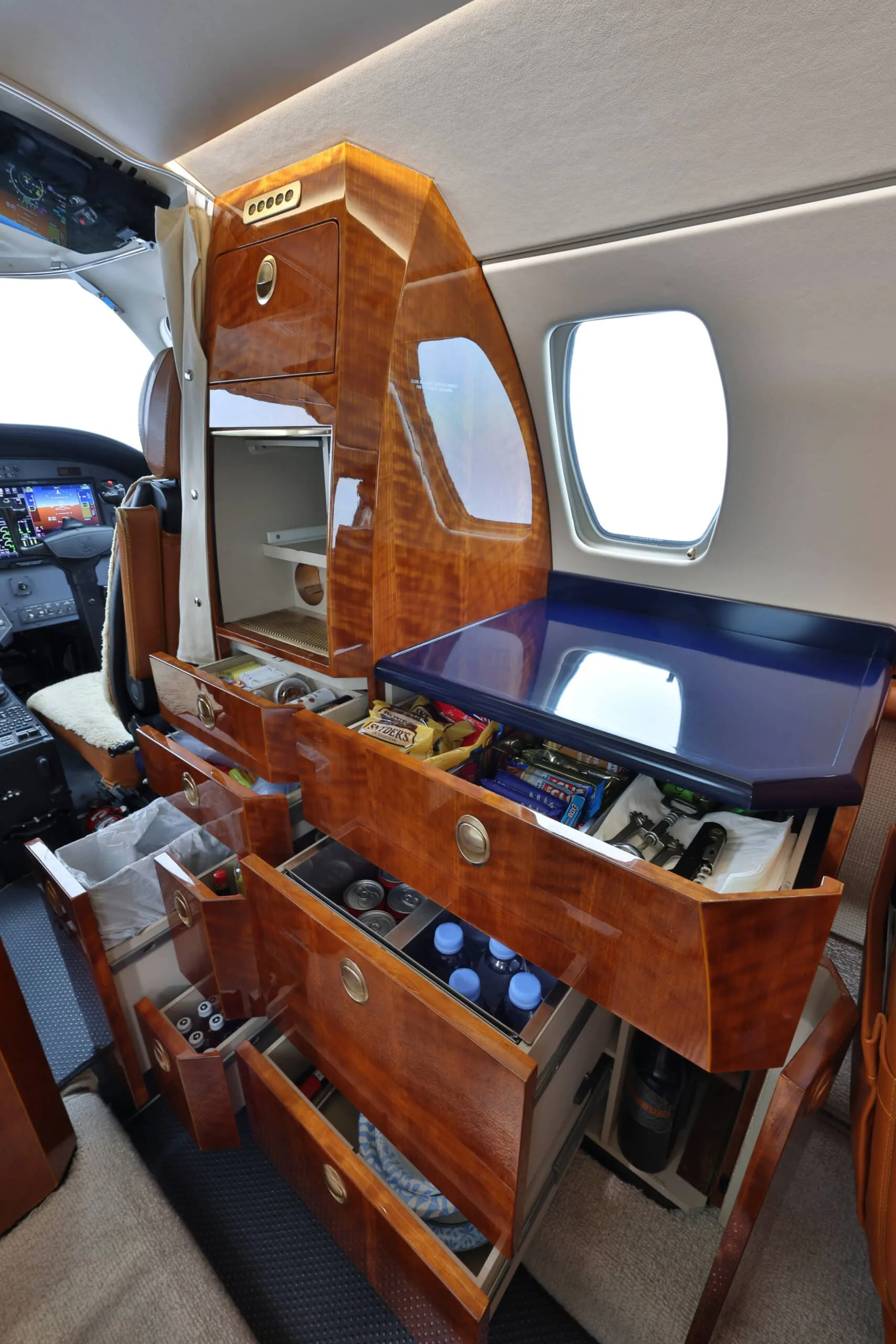 Open drawers of a private jet's polished wooden galley cabinet revealing snacks and drinks.