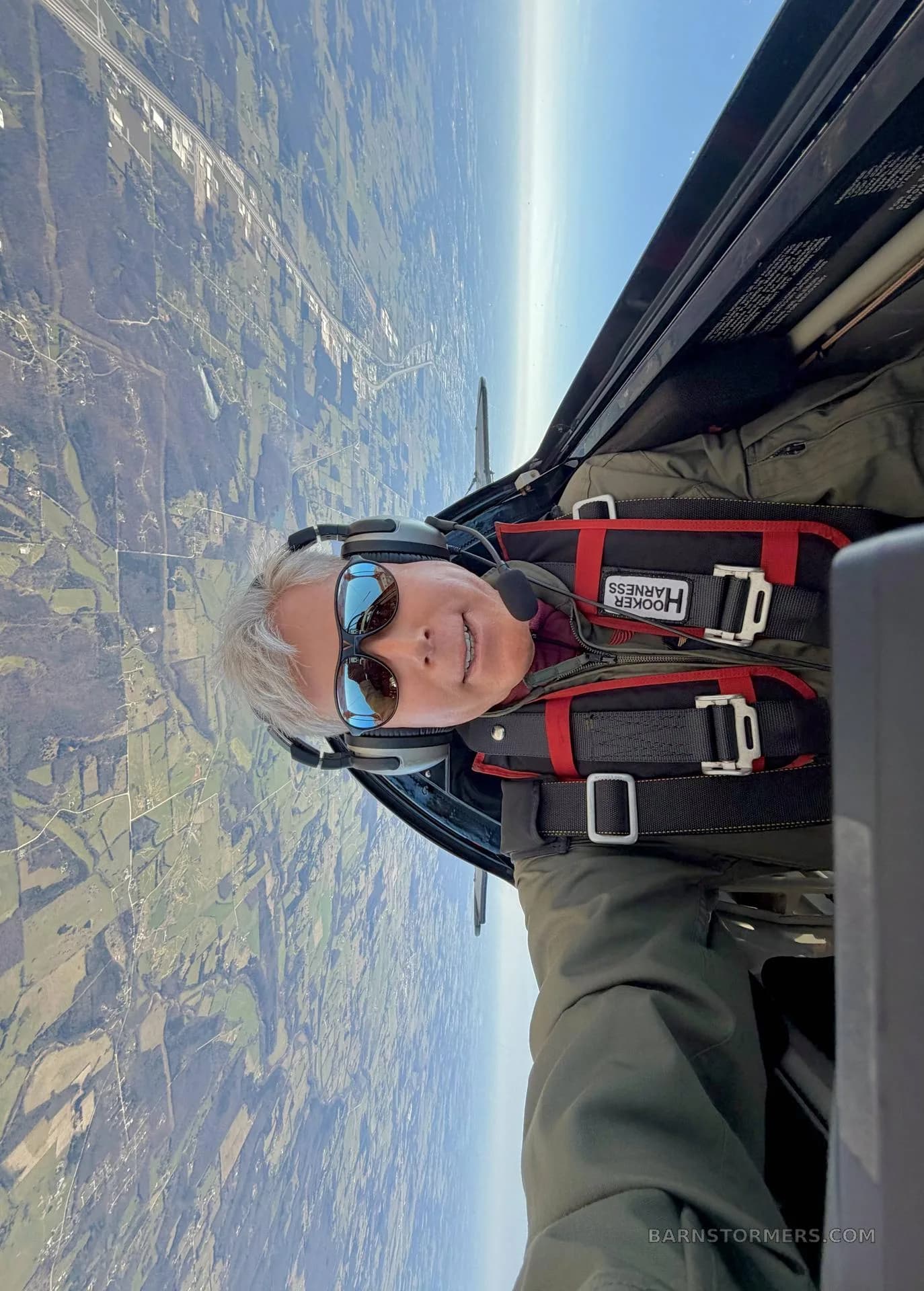 A pilot in a Pitts biplane cockpit, wearing a harness and sunglasses, looking at the camera while flying inverted over a green landscape.