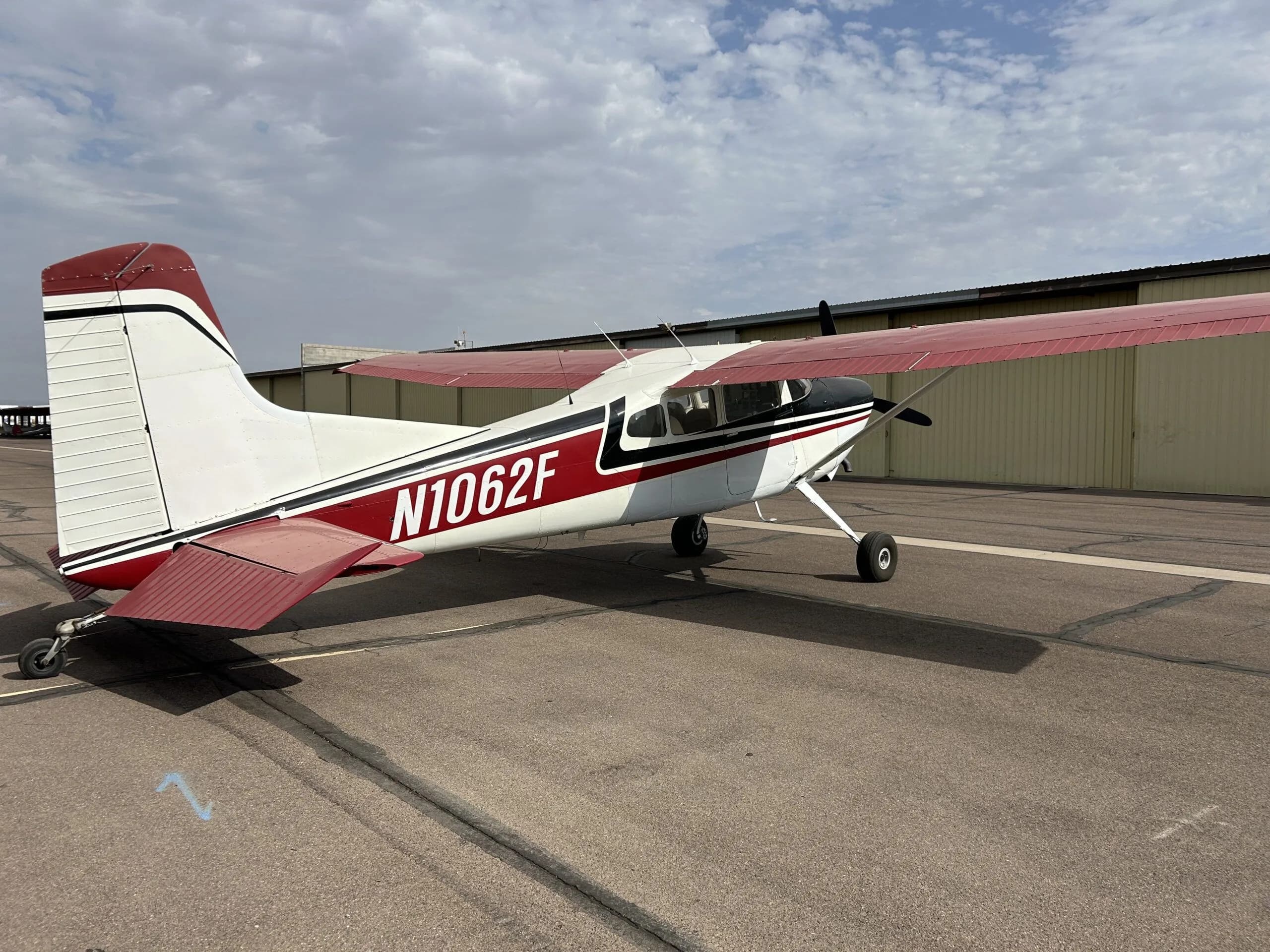 A white, black, and red Cessna 180 aircraft (N1062F) parked on an airport tarmac, viewed from the rear-right.