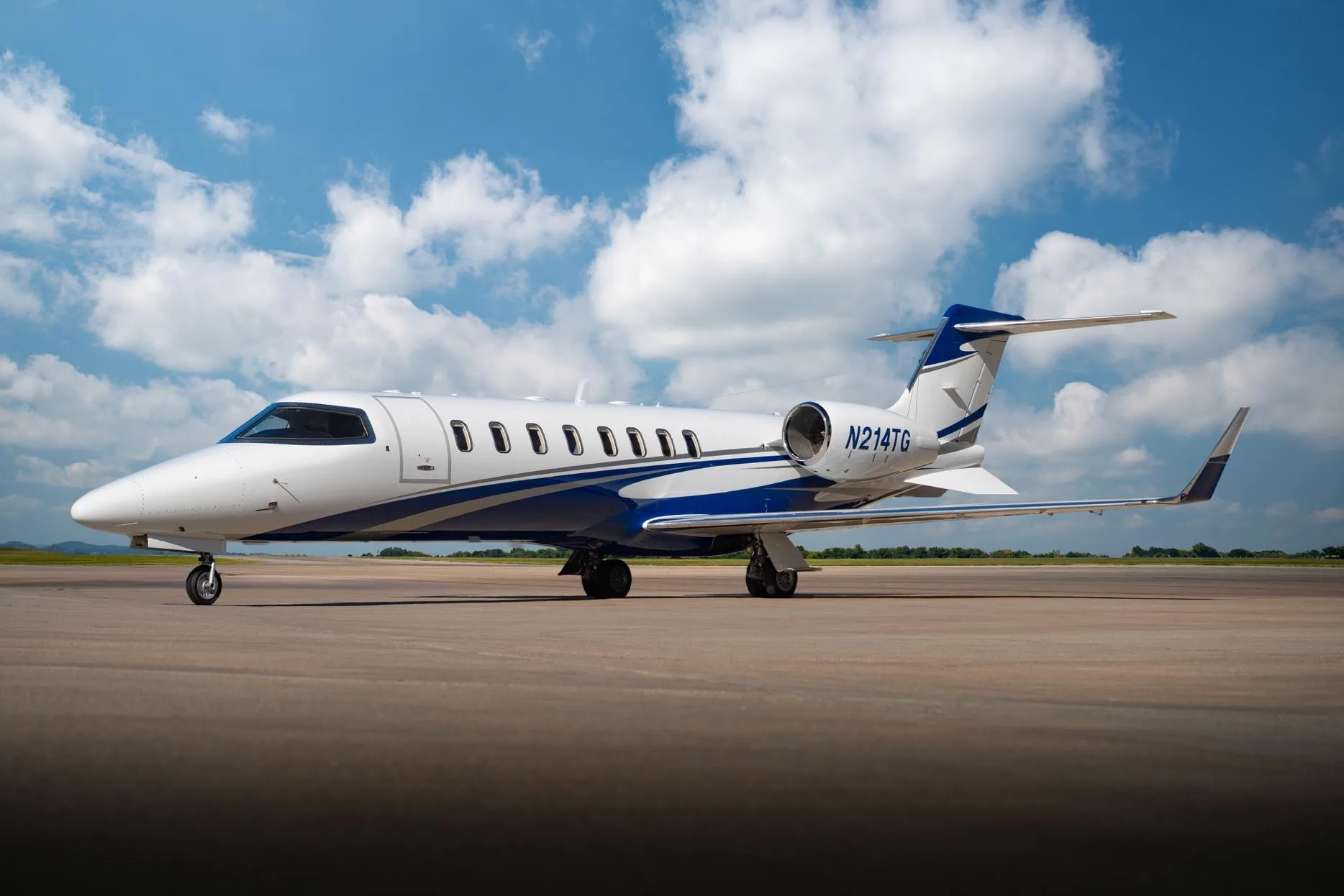 Side view of a white and blue Learjet 75 business jet, registration N214TG, parked on a tarmac under a partly cloudy sky.