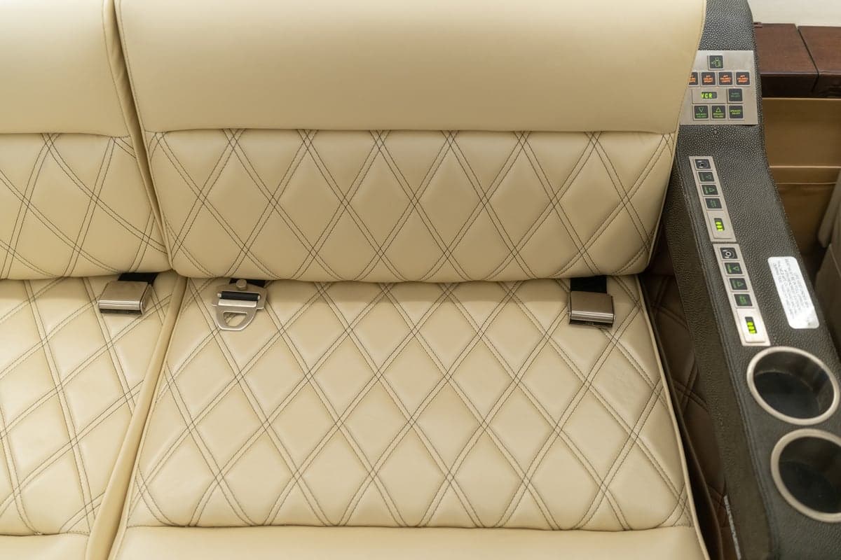 Close-up of cream-colored leather aircraft seats with diamond stitching and seatbelts, next to an armrest with controls and cup holders.