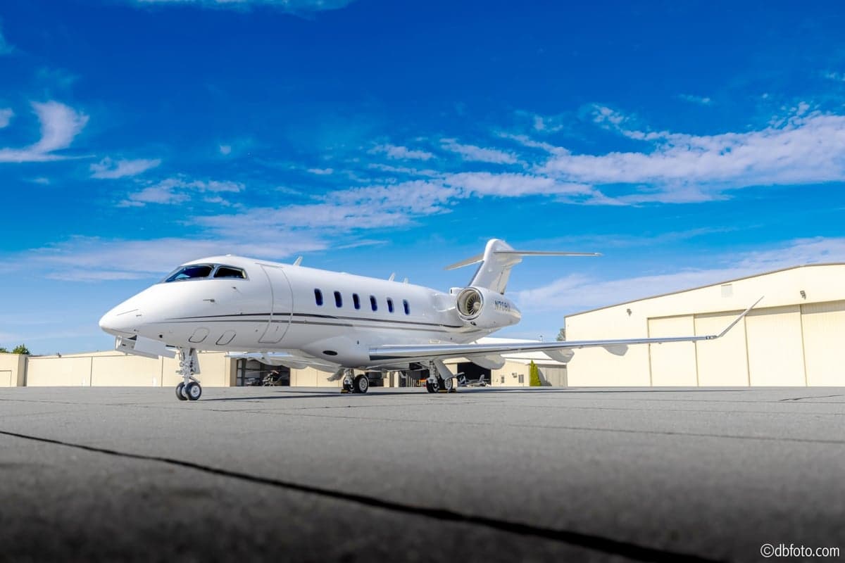 A white Bombardier Challenger business jet is parked on an airport tarmac with hangars in the background under a blue sky.