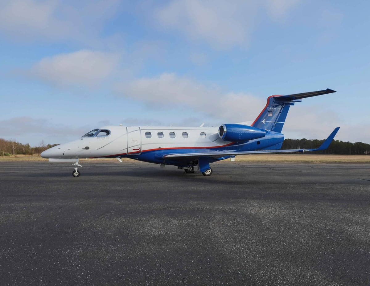 A Phenom 300E aircraft is parked on the tarmac with a blue and white paint scheme.