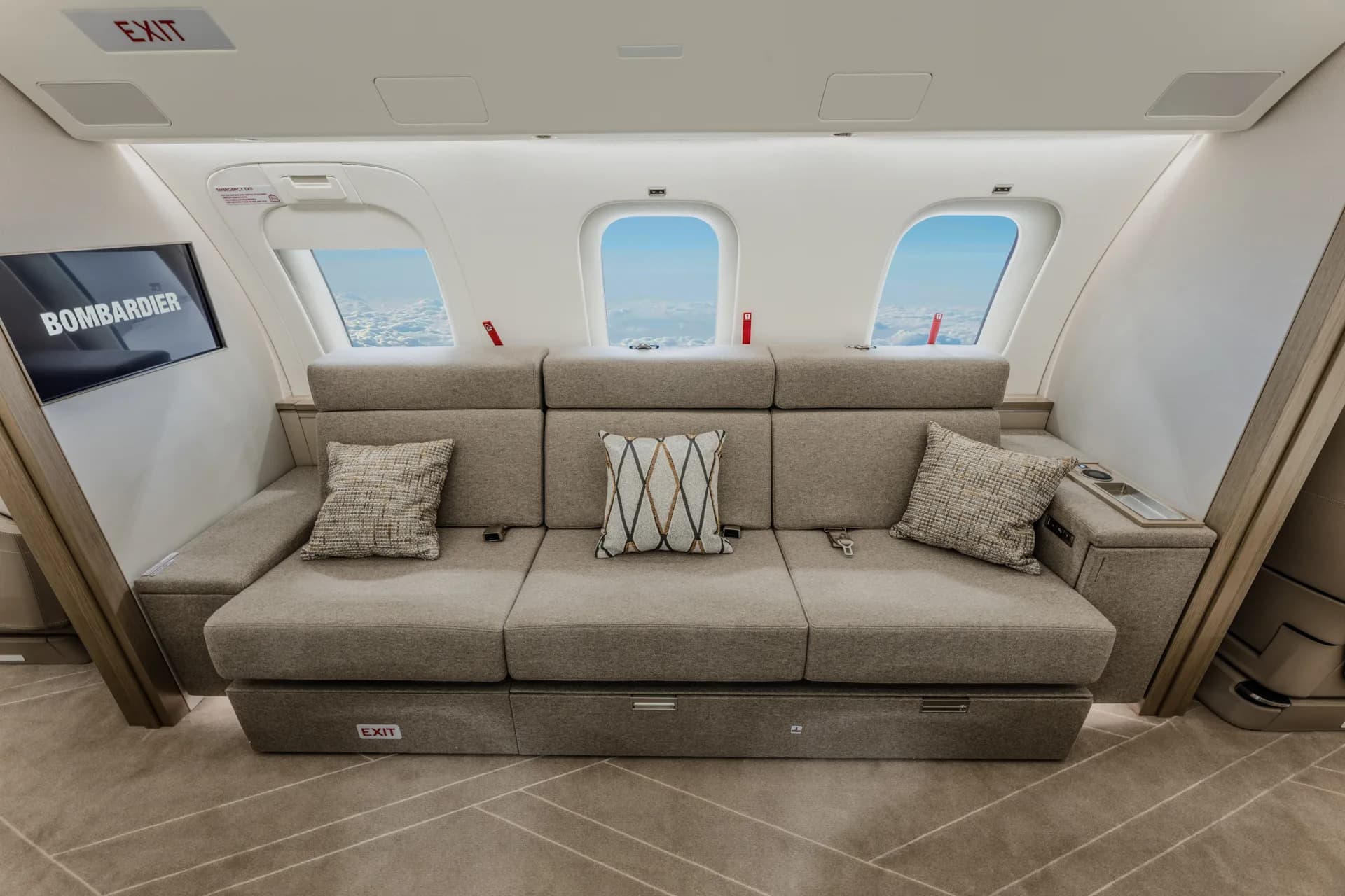 A close-up of a grey sofa with pillows inside a private jet cabin, with a Bombardier screen visible.