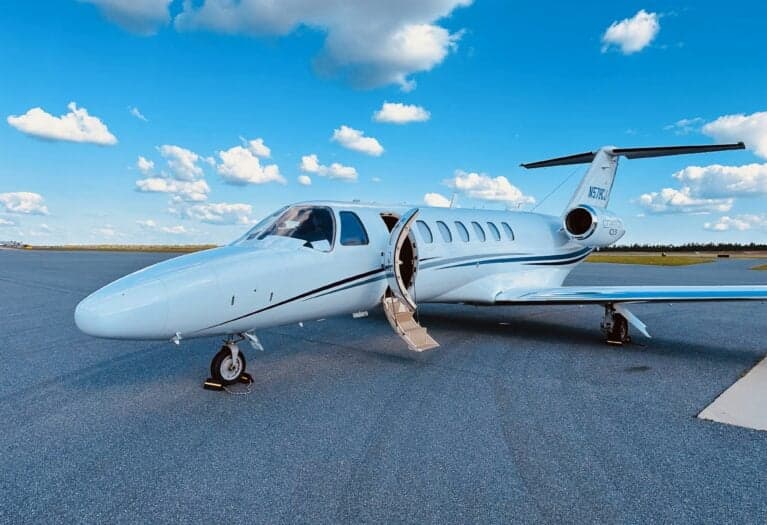 A white Cessna Citation CJ3 (N578CJ) with blue and grey stripes, parked on an asphalt tarmac with its main door open, under a blue sky with white clouds.