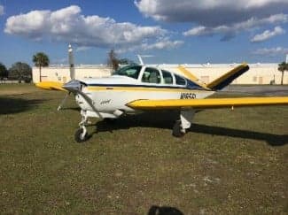 A Beechcraft Bonanza aircraft with a white, yellow, and blue paint scheme is parked on the grass.