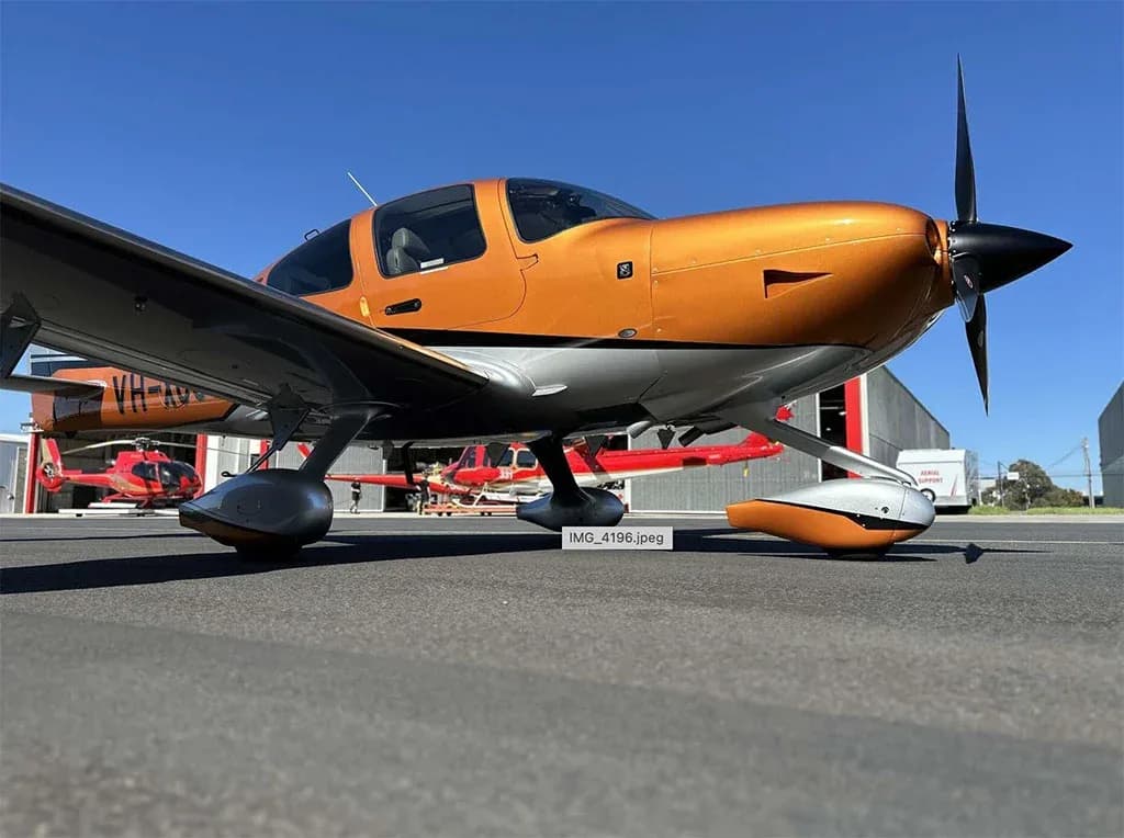 A low-angle front view of an orange and silver Cirrus SR22 aircraft with registration VH-XC6 on an airport tarmac, with other aircraft in the background.