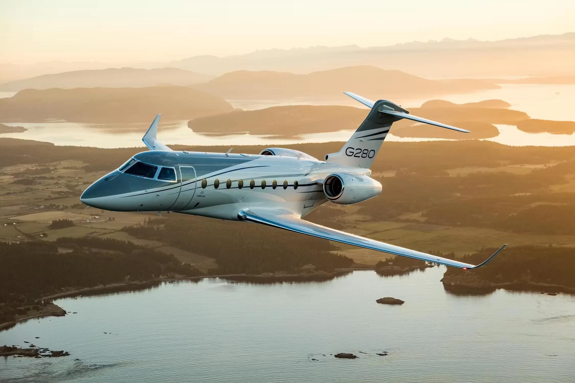 A white and blue Gulfstream G280 business jet flies over a scenic coastal landscape with islands and mountains at sunset.