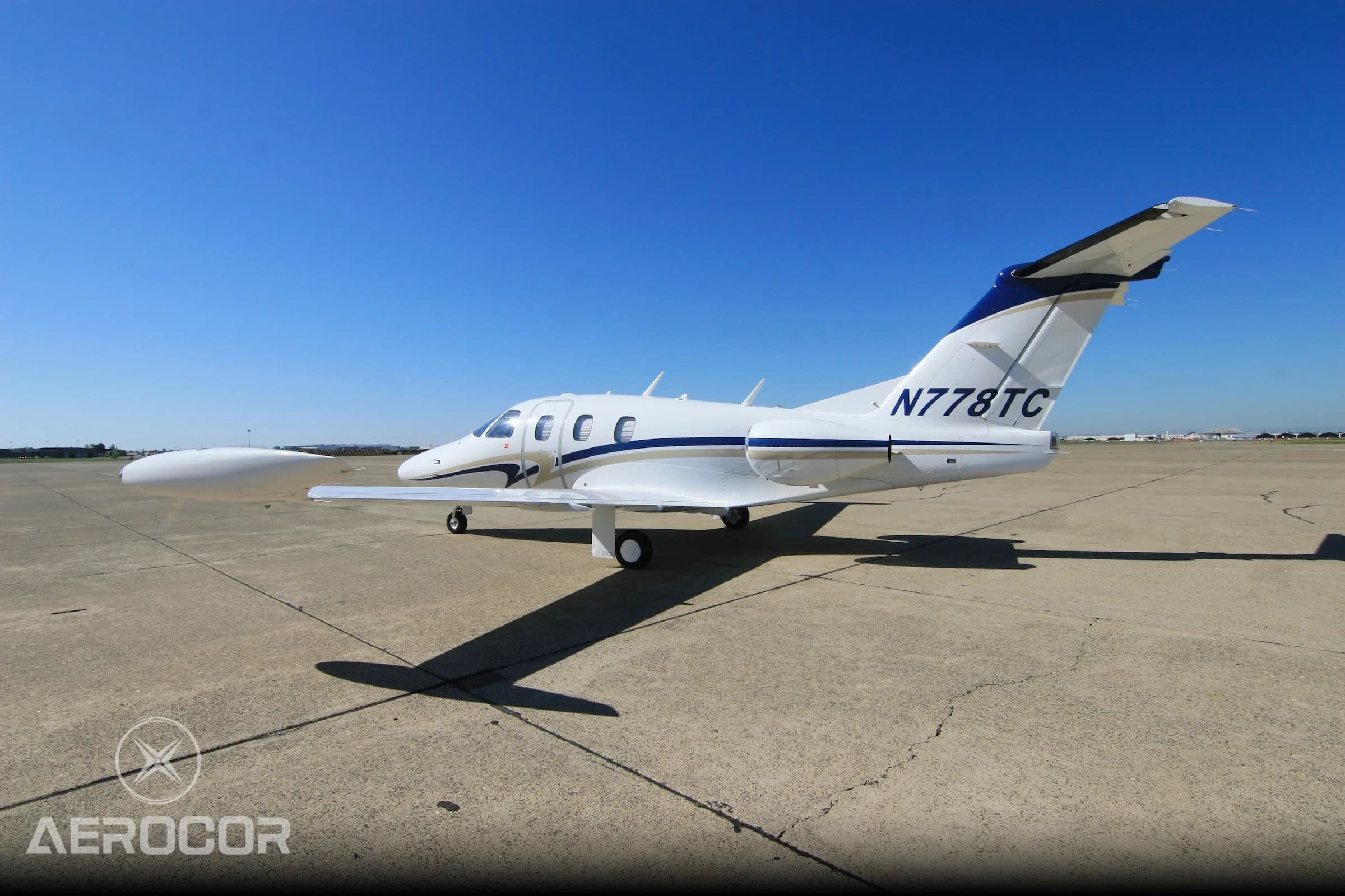 A white Eclipse 500 jet with blue and gold stripes, viewed from the rear-right on the tarmac.