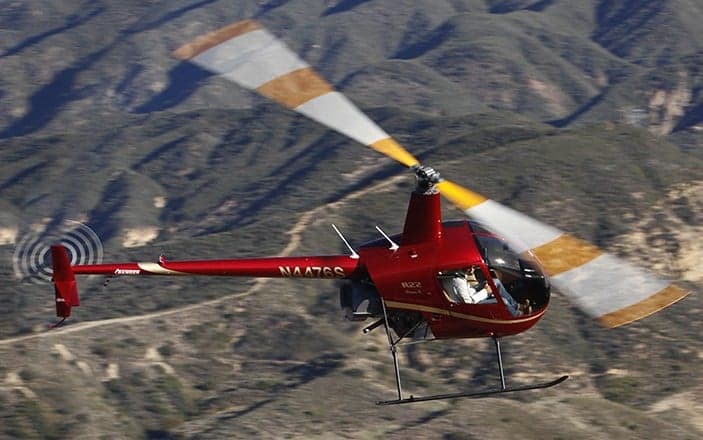 A red Robinson R22 helicopter flying over a mountainous area.