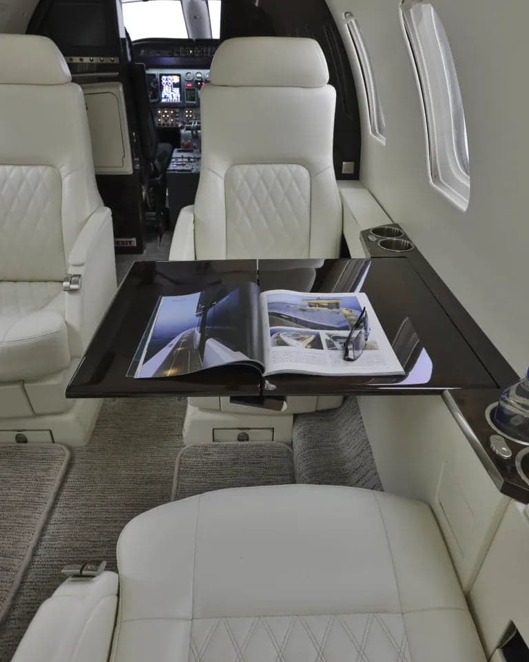 Aircraft interior with white leather seats, a dark wood table with a magazine, and a view of the cockpit.