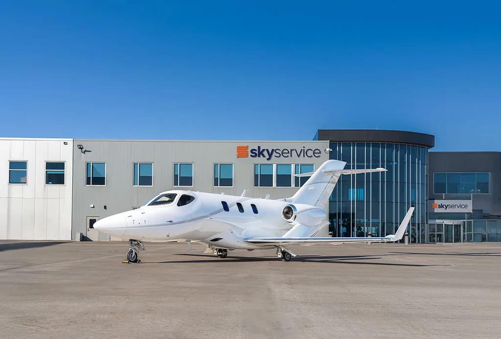 A white HondaJet HA-420 business jet parked on a tarmac in front of a "skyservice" hangar under a clear blue sky.