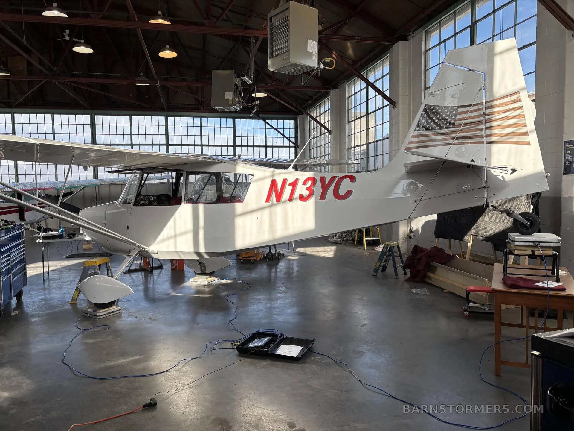 A light gray single-engine aircraft with red N13YC registration and an American flag on the tail, parked in a hangar.