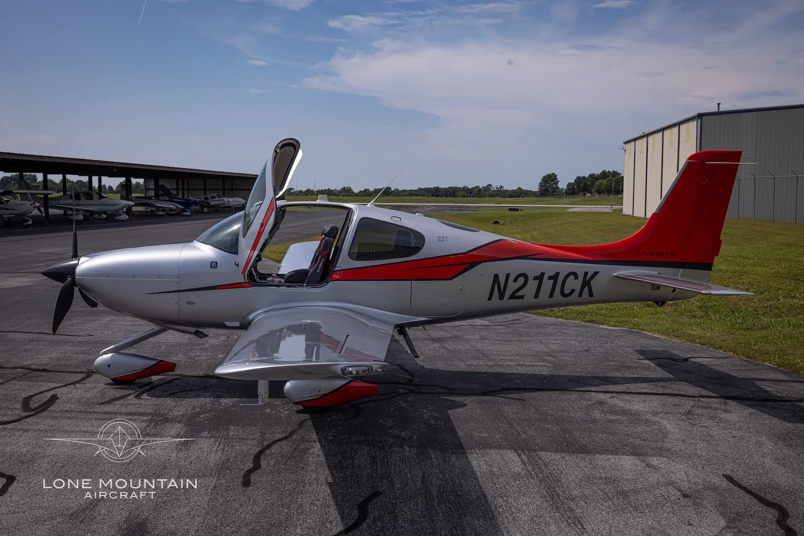 A side view of a silver and red Cirrus SR22T aircraft with registration N211CK parked on an asphalt ramp with its gull-wing doors open.