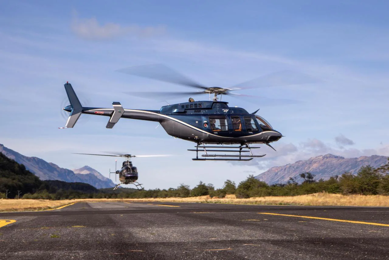 Two dark blue helicopters, possibly Bell 407s, taking off from a runway with mountains in the background.