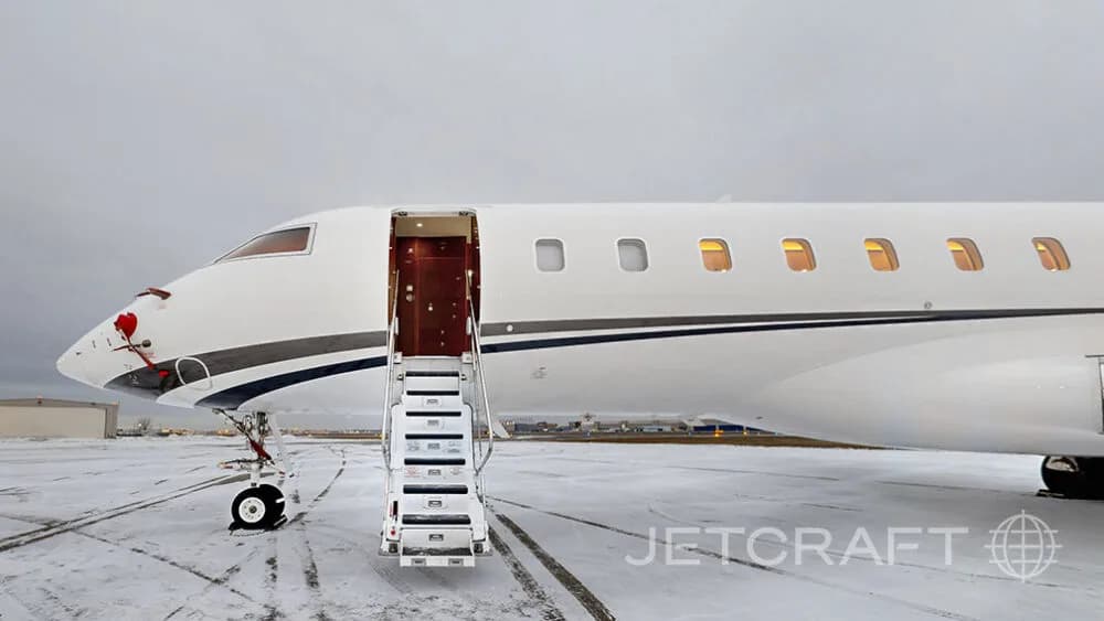 A close-up view of the front section of a white Bombardier Global Express business jet on a snowy tarmac, with its main door open and stairs extended.