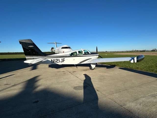 A Beechcraft Bonanza parked on the tarmac with a jet in the background.