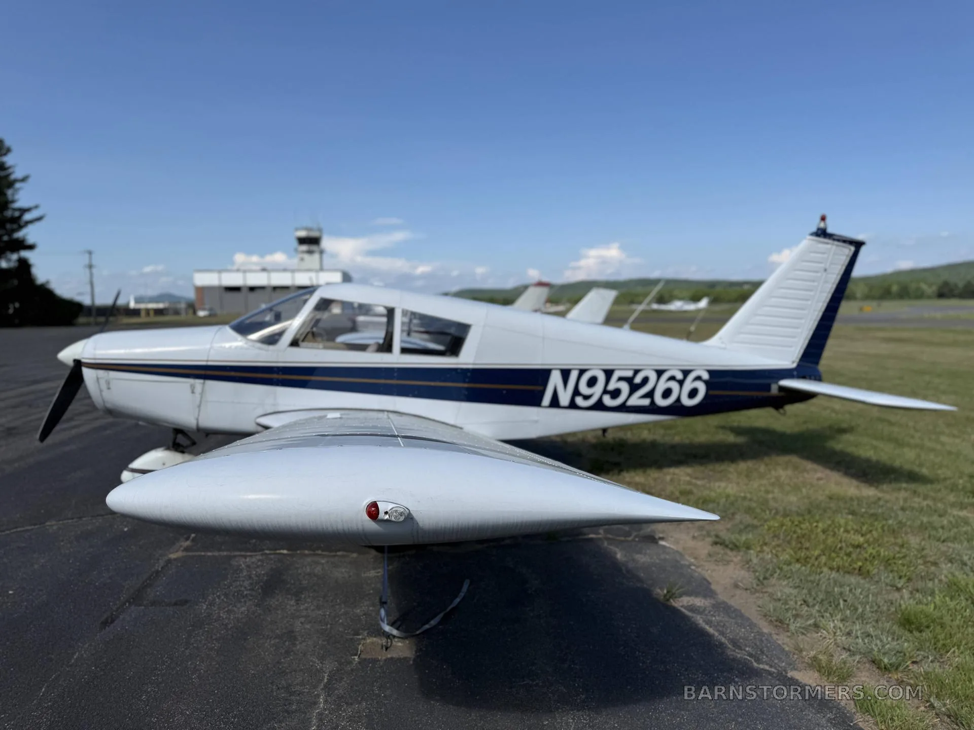A white single-engine propeller aircraft (N95266) with blue stripes, viewed from the side, showing the wing and fuselage with an air traffic control tower in the background.