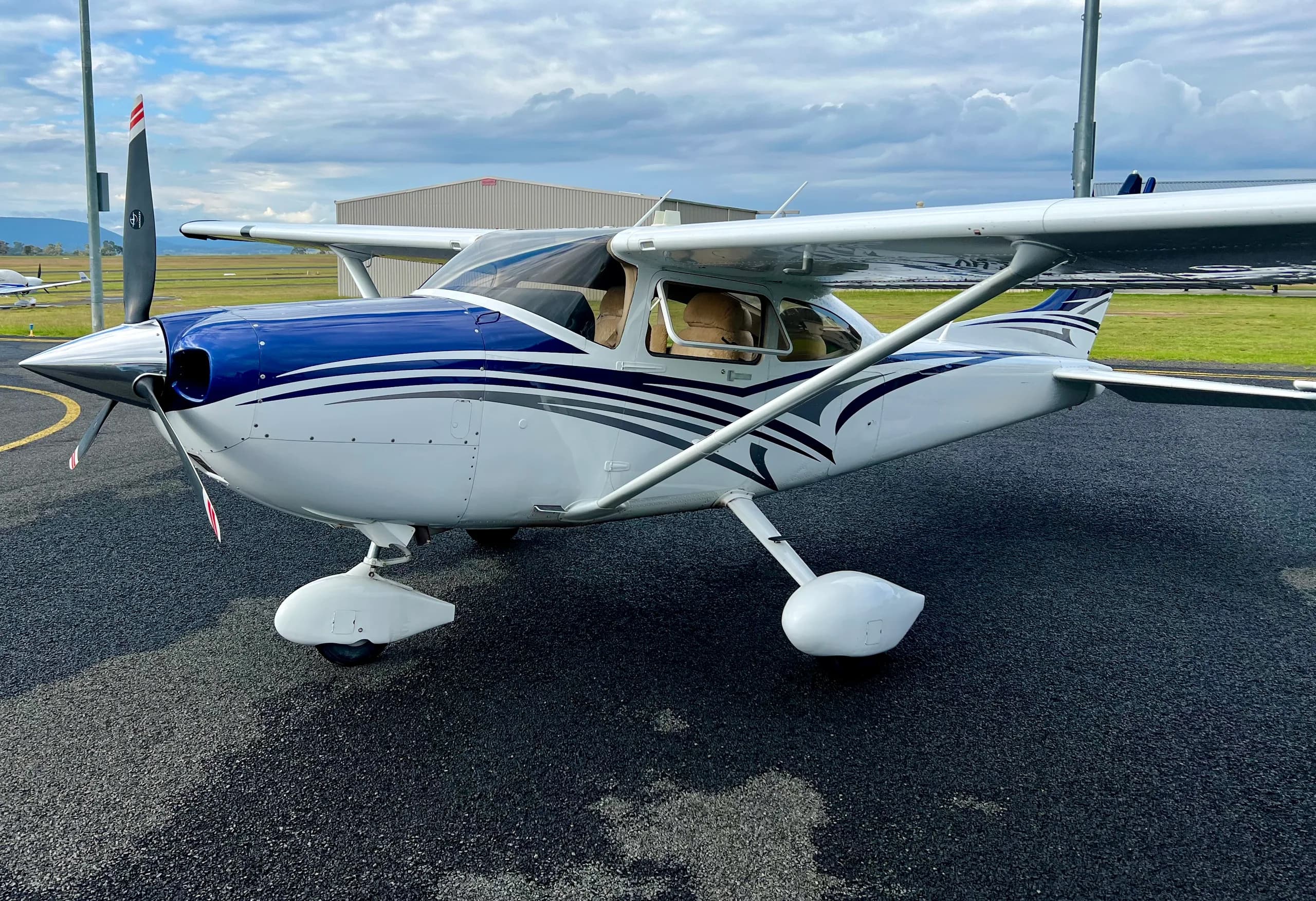 A white single-engine, high-wing propeller aircraft with blue and grey stripes is parked on an asphalt apron at an airfield.