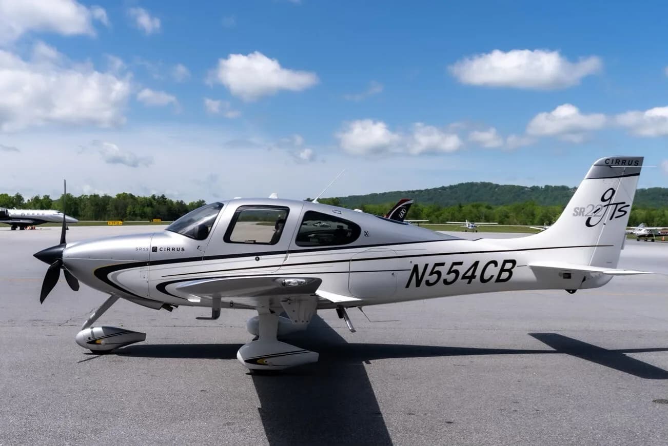 A side view of a silver and white Cirrus SR22T aircraft with registration N554CB parked on an airport tarmac under a blue sky.