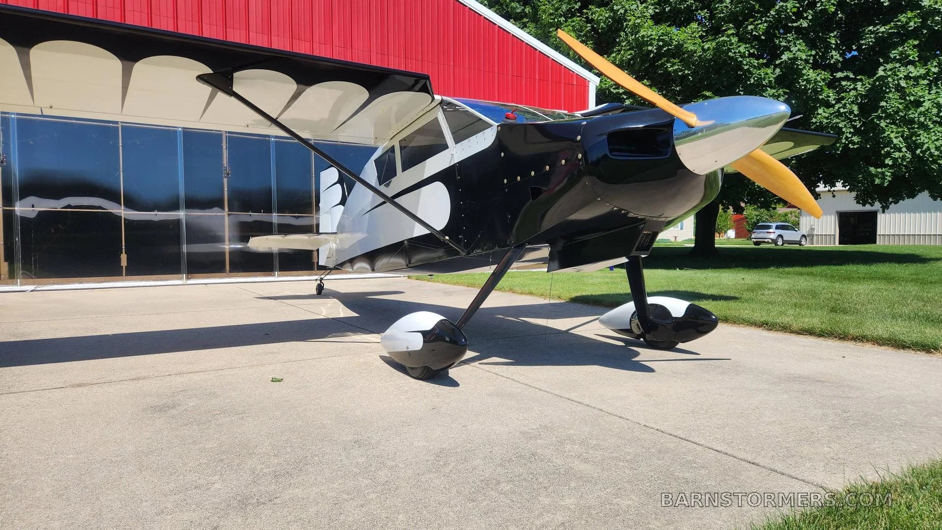 A black and white single-engine aircraft with a wooden propeller is parked on concrete, viewed from the front, next to a red hangar.