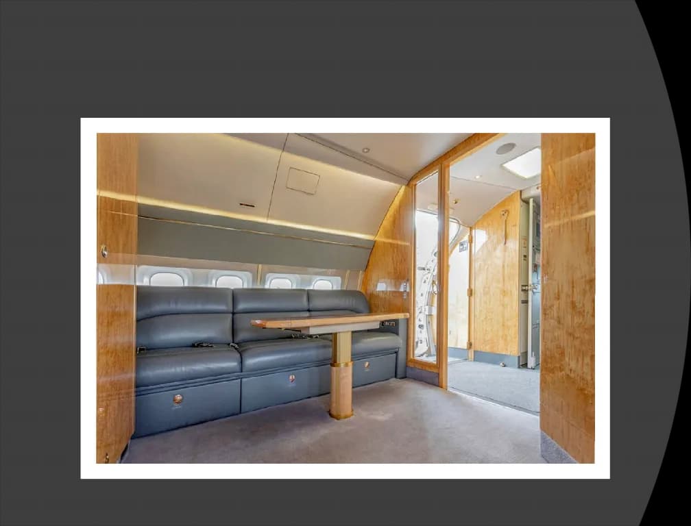 A private jet cabin interior with a dark grey leather sofa, a wooden table, and light wood paneling.