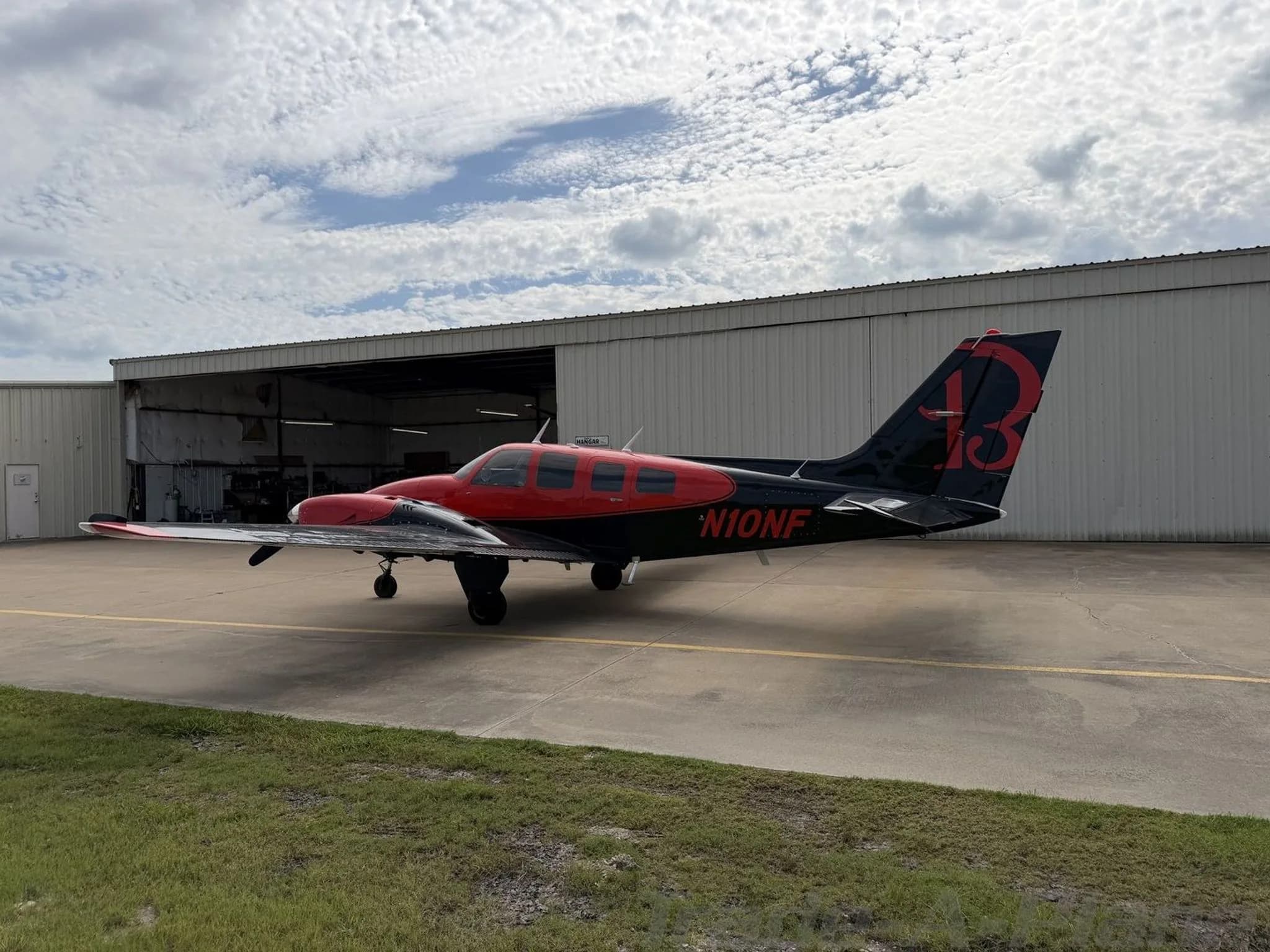 Side view of the red and black twin-engine propeller aircraft, N10NF, with a large red "13" on its tail, parked in front of an open hangar.