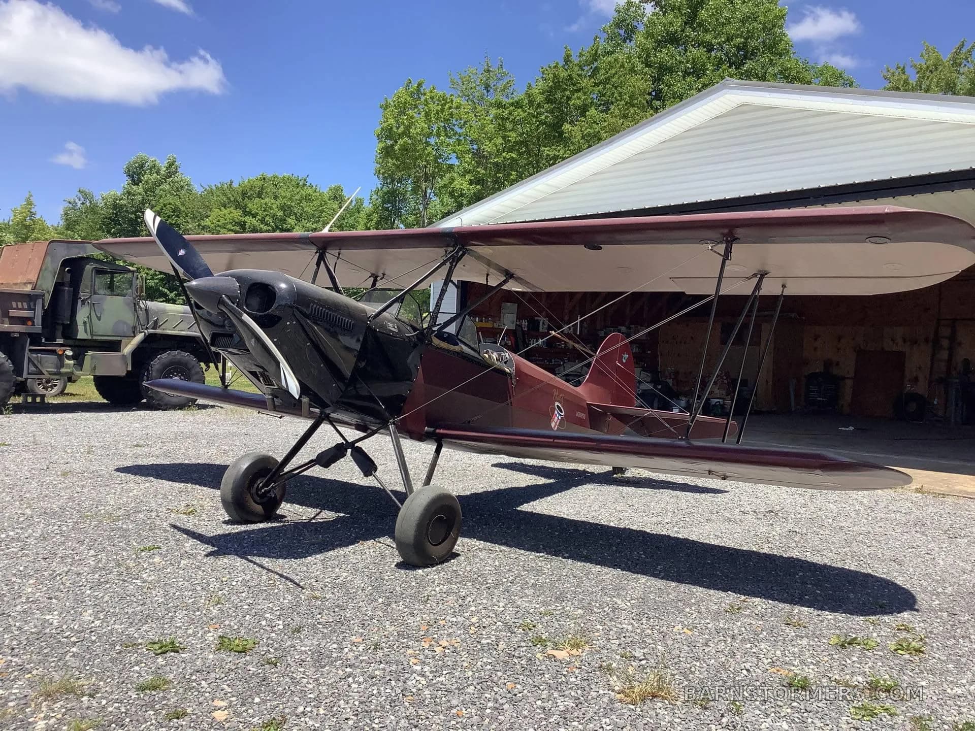 A maroon and black biplane with a black engine cowling, parked on gravel next to a hangar and a green truck.