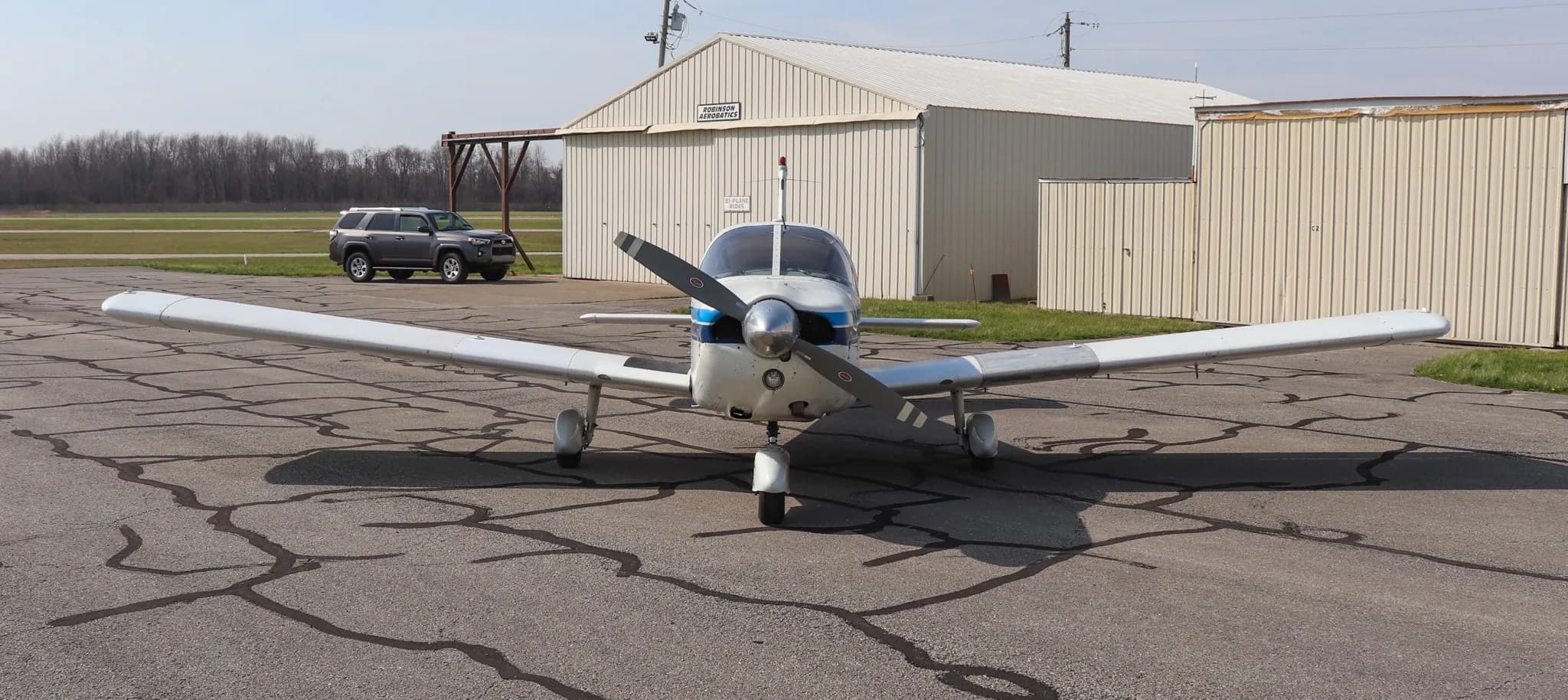 A front view of the white Piper Cherokee 235 aircraft with blue stripes, parked on an asphalt ramp with hangars in the background.