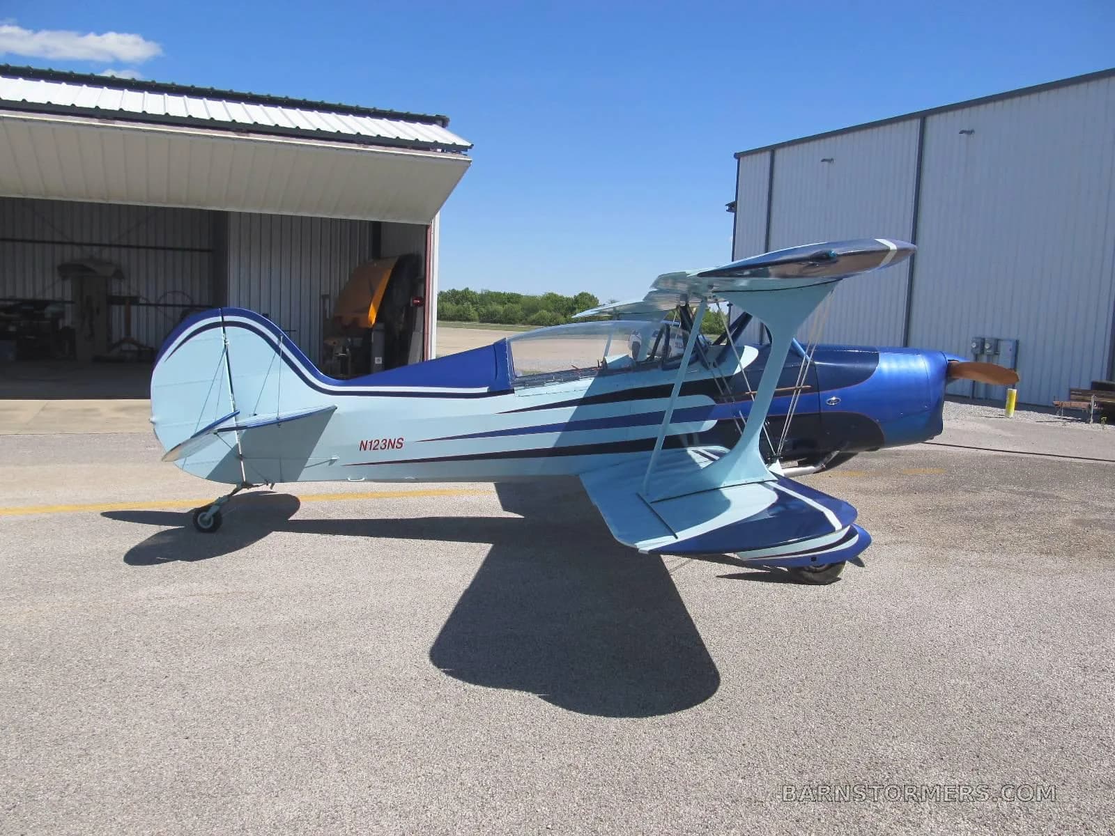 A blue and light blue biplane with an orange propeller spinner is parked on an asphalt apron between two hangars.