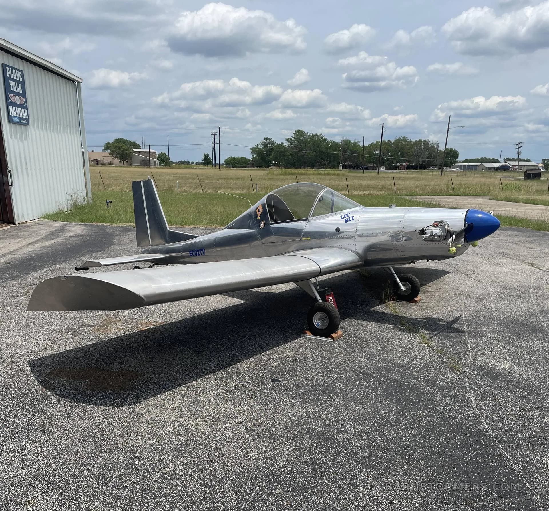 A silver single-engine aircraft with a blue nose cone is parked on an asphalt apron next to a hangar under a cloudy sky.