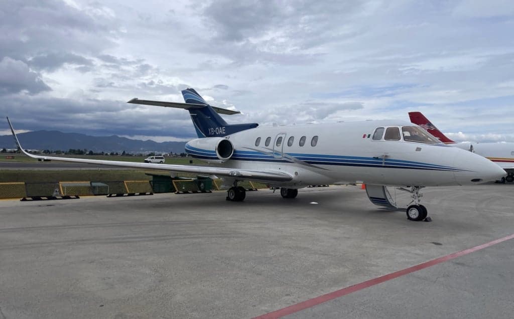 A white business jet with blue stripes (XB-OAE) parked on a tarmac under a cloudy sky, with mountains in the background.